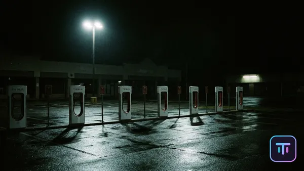 A desolate Tesla Supercharger station at night in a rain-slicked suburban parking lot, charging stalls standing empty under a single harsh floodlight with closed storefronts in the background