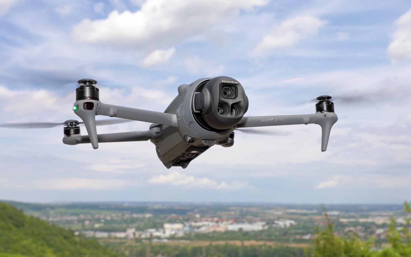 DJI Mavic 4 Pro in flight against a partly cloudy sky, triple-lens Hasselblad gimbal facing the camera