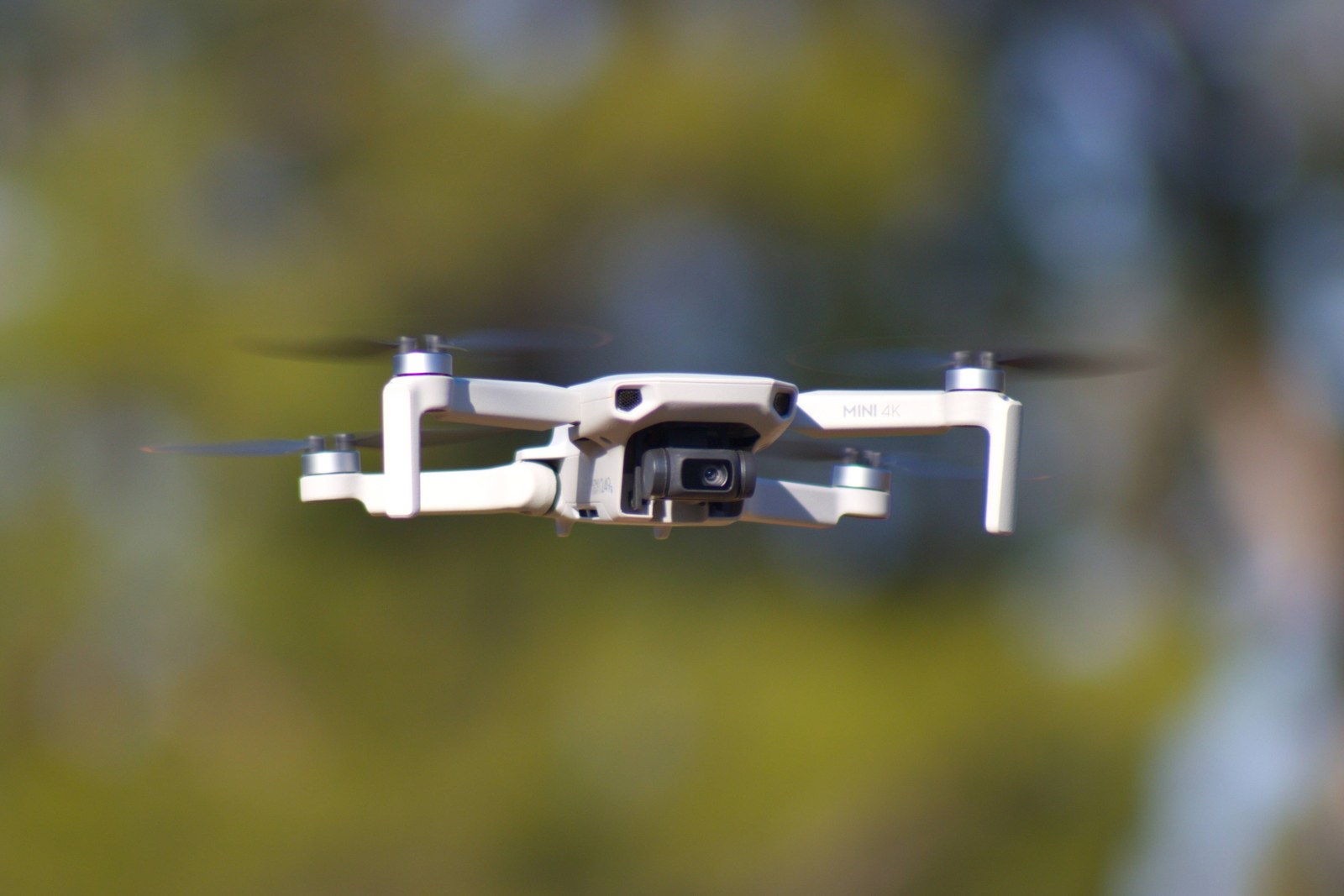 DJI Mini 4K drone hovering in front of blurred green trees, MINI 4K lettering visible on its arm