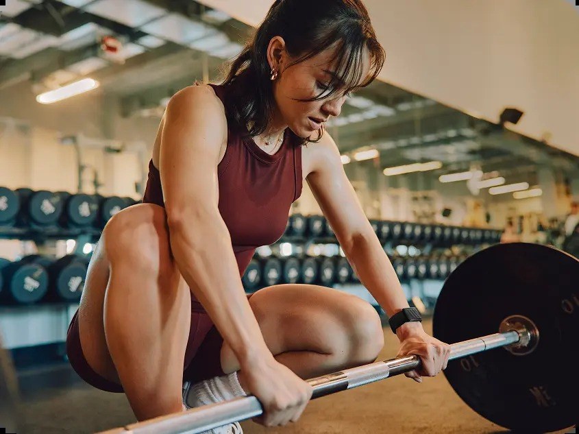 A woman lifting a barbell in a gym wearing a WHOOP 5.0 band on her wrist