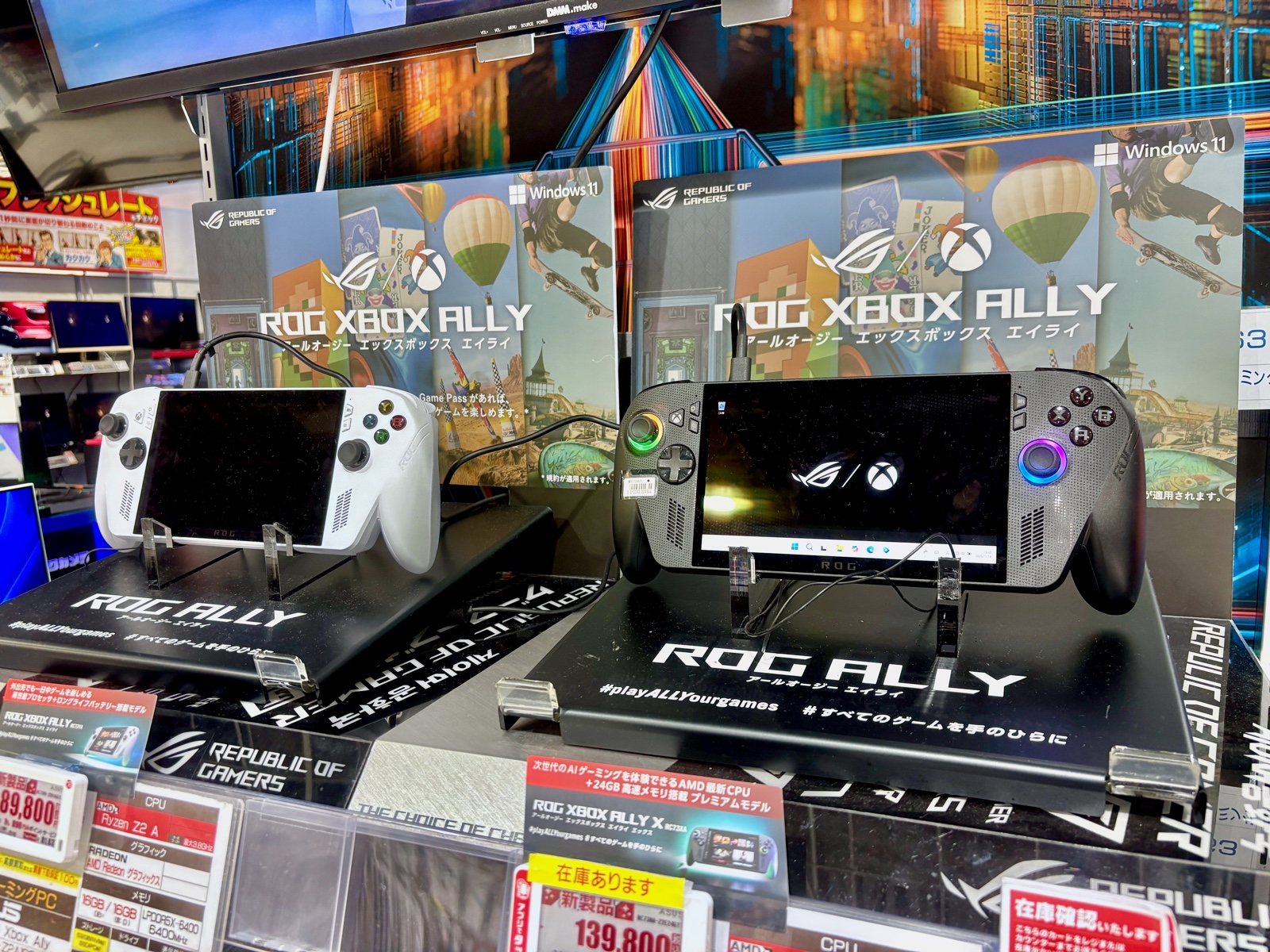 The white ROG Xbox Ally and black ROG Xbox Ally X handhelds on a retail display stand under Windows 11 signage