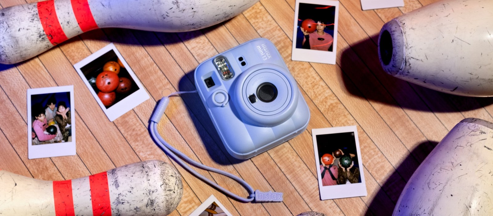 A Frost Blue instax mini 13 instant camera lying on a bowling lane surrounded by credit-card-sized prints and bowling pins