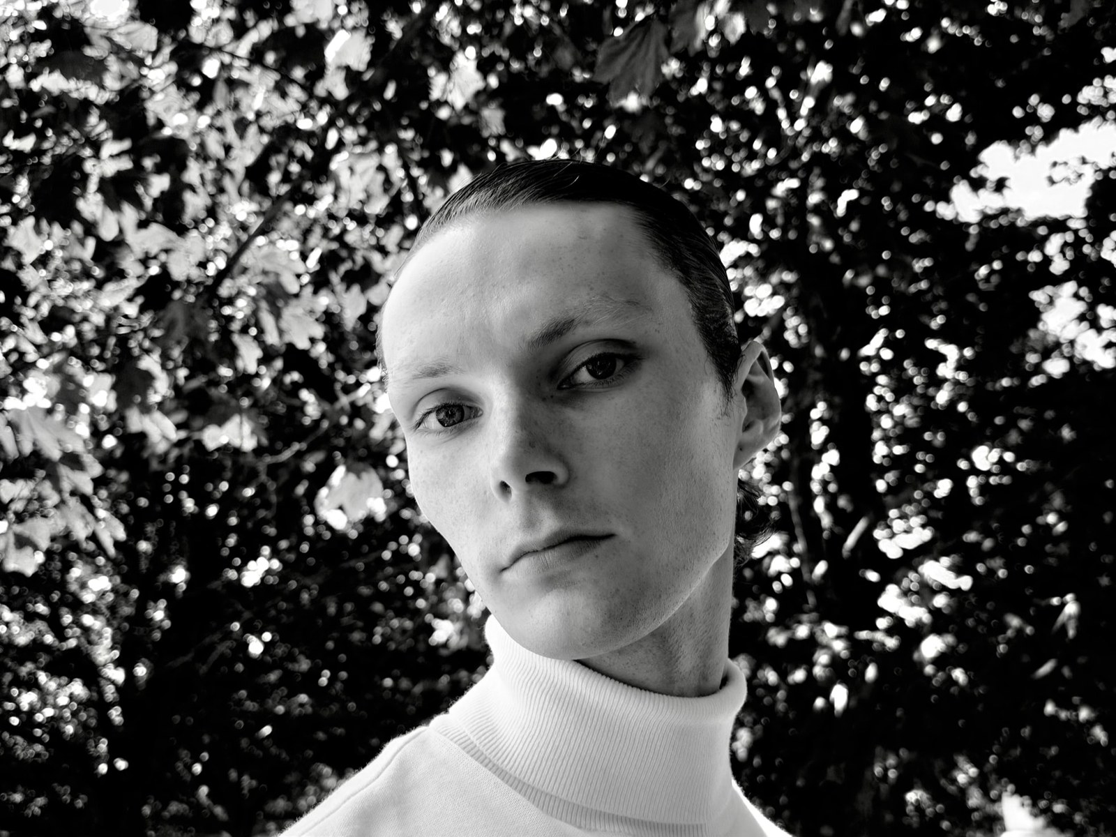 A black-and-white portrait of a young man in a turtleneck against foliage, shot on iPhone 18 Pro with the variable aperture set for shallow depth of field