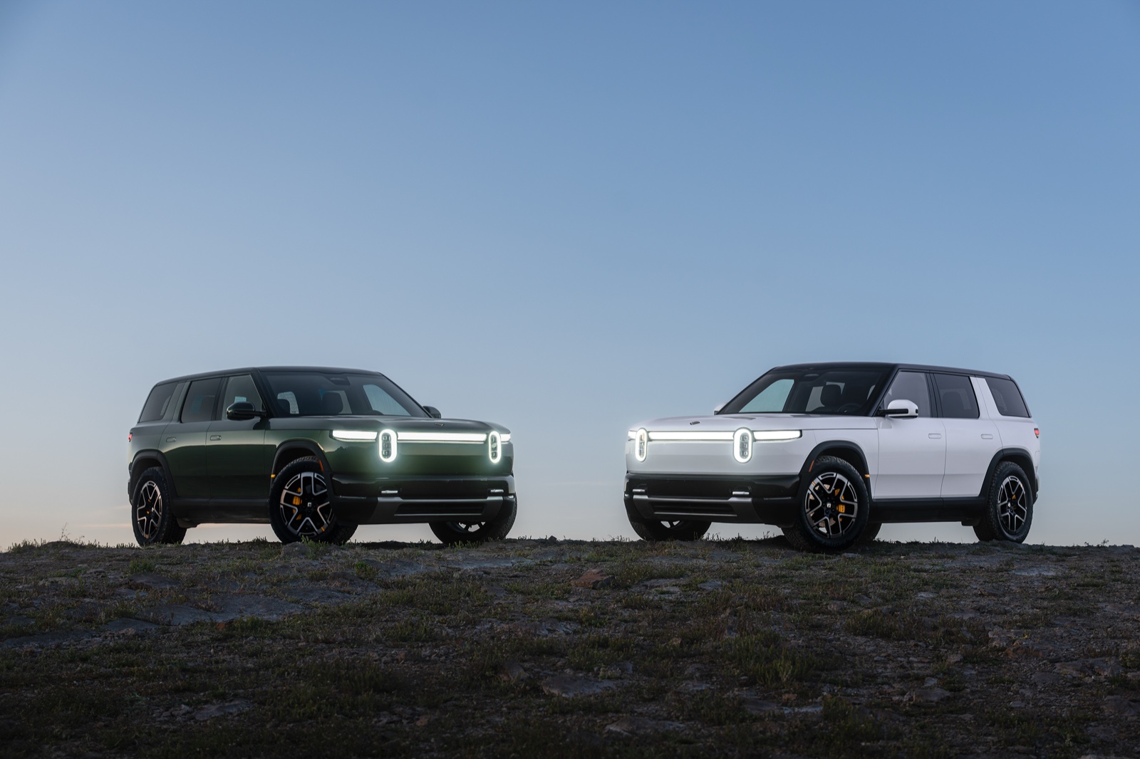 Two Rivian R2 electric SUVs in green and white parked side by side on a rocky hilltop at dusk