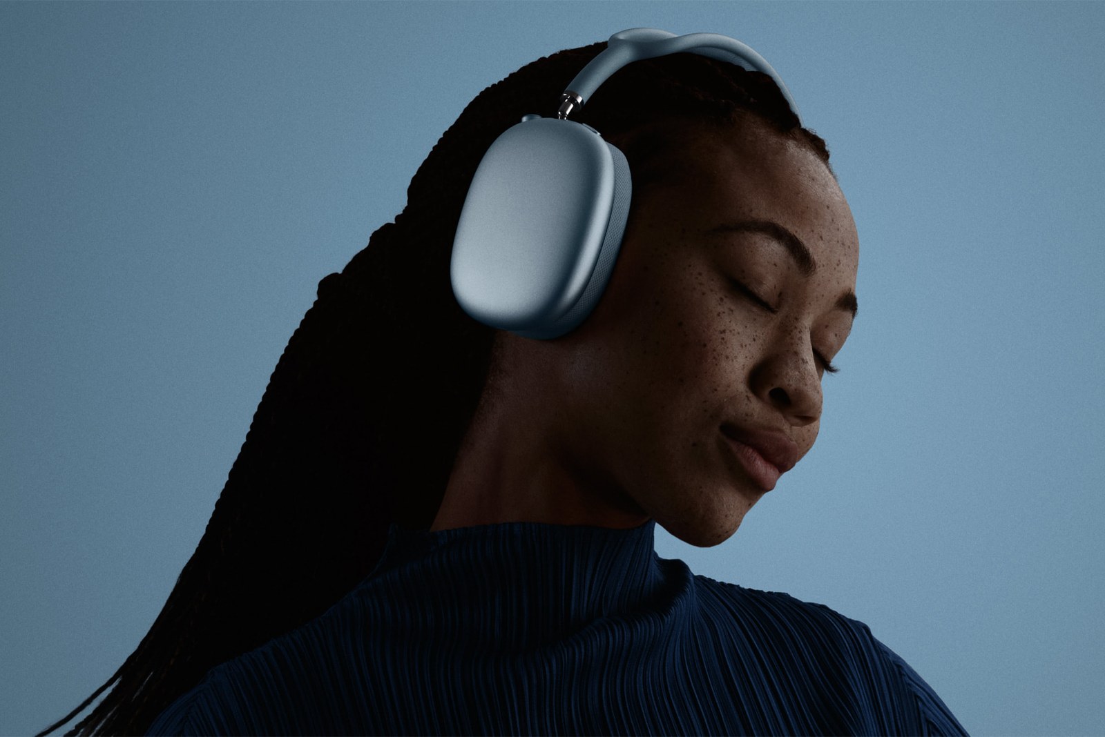 A woman with her eyes closed wears light blue AirPods Max 2 over-ear headphones against a blue background