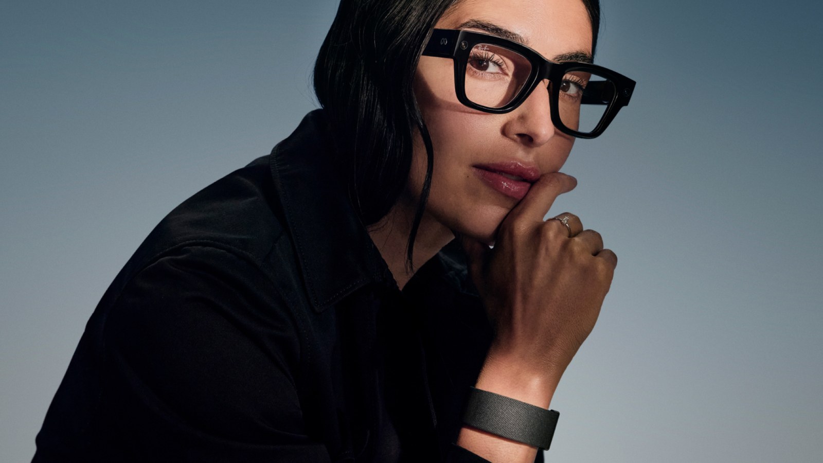 A woman in thick black Meta Ray-Ban Display frames rests her chin on her hand, with the black Meta Neural Band on her wrist